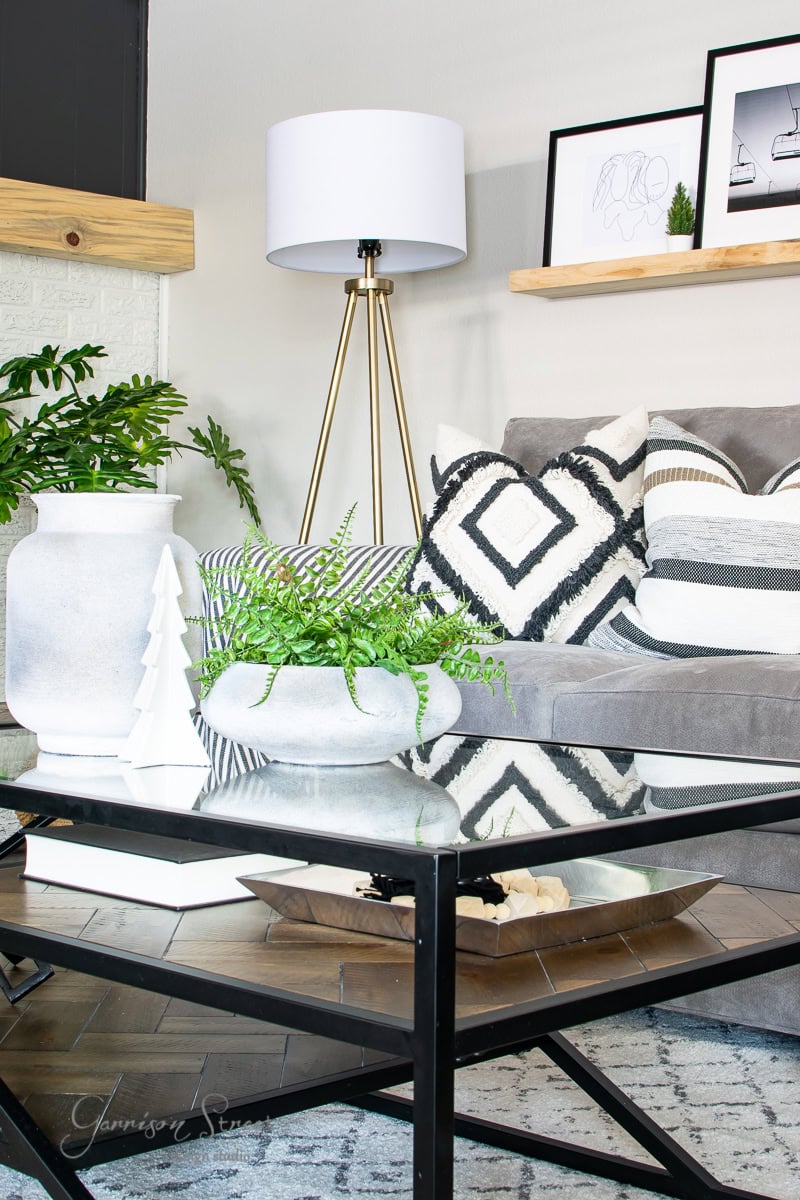 A modern living room from the 2020 Christmas house tour features a gray sofa adorned with patterned pillows. A glass coffee table holds a potted fern, while a brass floor lamp with a white shade stands nearby. Minimalist art decorates the wall above wooden shelves, enhancing the festive charm.