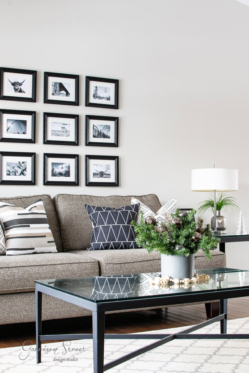 Modern living room with a gray sofa adorned with black and white patterned pillows, reminiscent of a 2020 Christmas house tour. A glass coffee table displays green foliage in a planter, while the wall features a grid of framed black and white photos. A sleek lamp graces the side table.