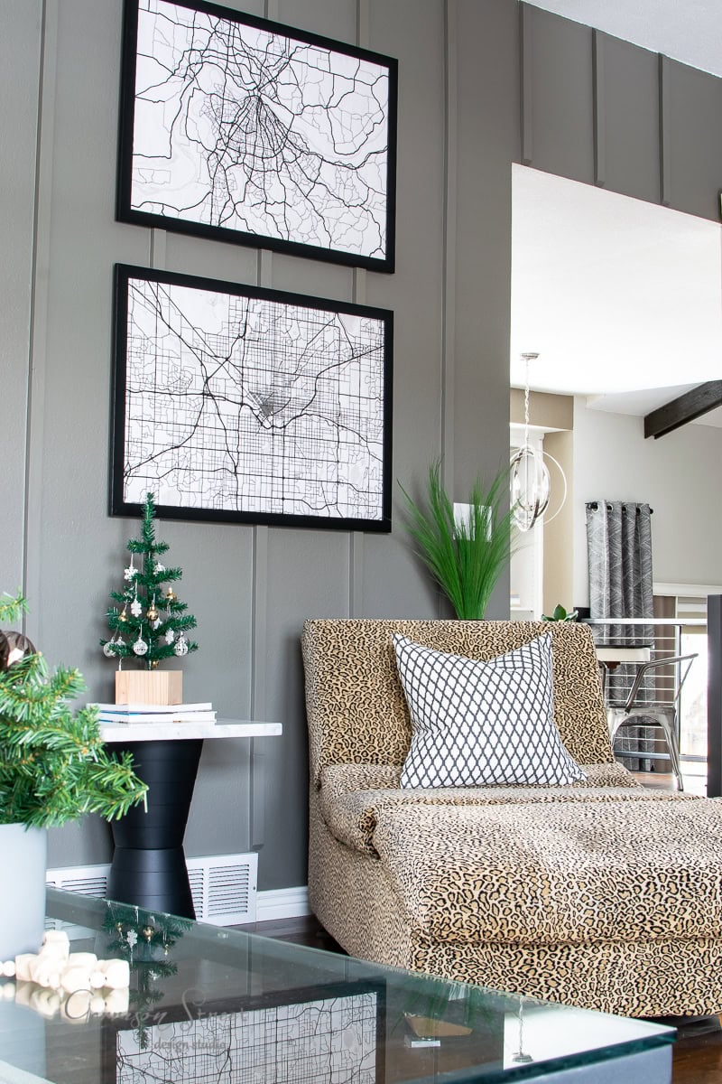 A living room with a modern design showcases a plush leopard-print chair adorned with a patterned pillow. Two framed city map prints grace the gray wall. A small tabletop Christmas tree adds festive charm to the white side table, hinting at a delightful 2020 Christmas house tour.