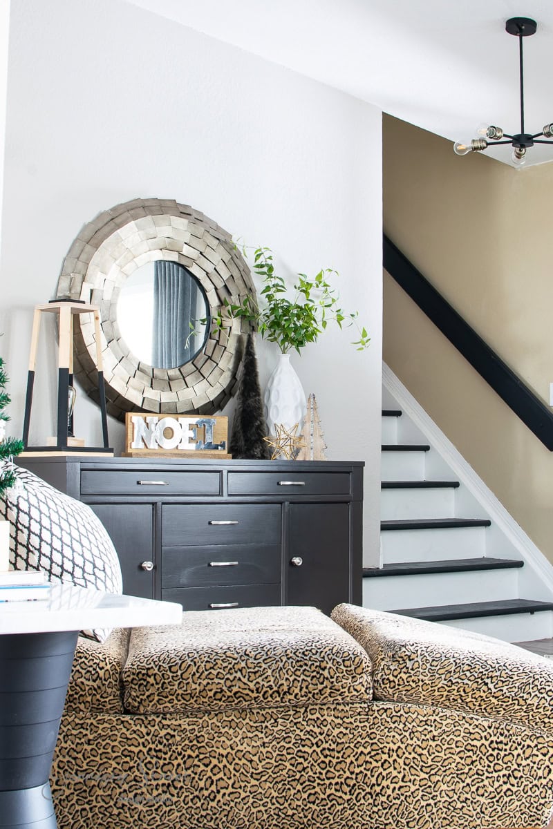 A stylish living room from a 2020 Christmas house tour features a dark dresser with a circular mirror above it, decorated with a small tree branch and "NOEL" letters. The room includes leopard-print furniture and a staircase with white steps and a black railing, topped by a modern light fixture.