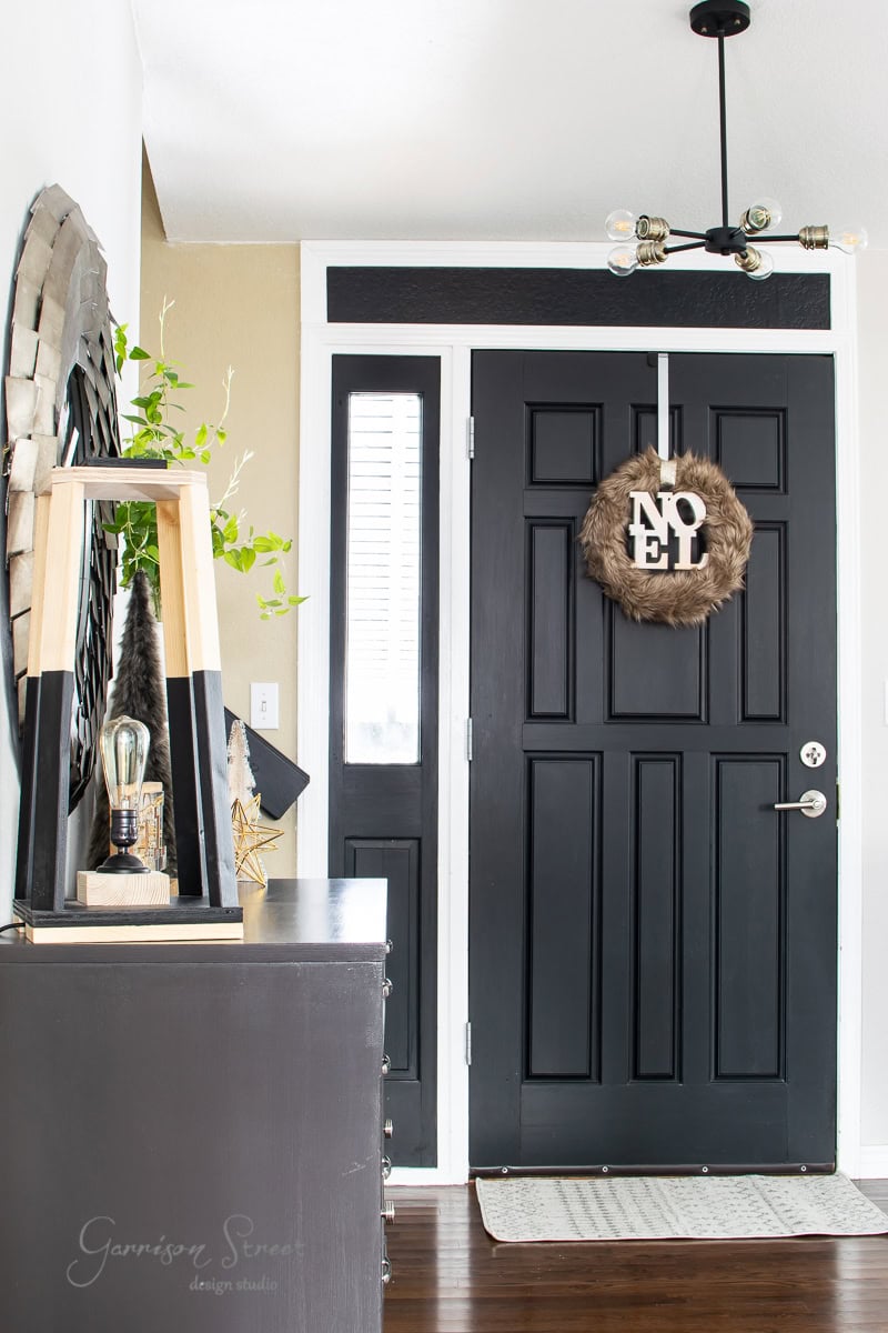Step into a festive 2020 scene with this modern entryway, featuring a black door adorned with a fur wreath and "Noel" sign. A sleek black dresser displays a lamp and vase of greenery, while an overhead fixture with exposed bulbs adds glow. A woven-framed wall mirror completes the Christmas house tour charm.
