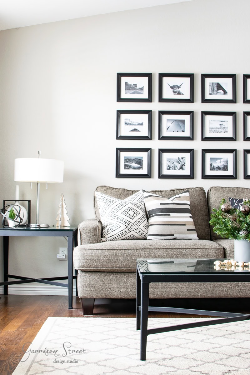 A modern living room, perfect for a 2020 Christmas house tour, features a gray sofa with decorative pillows, a black coffee table, and a side table with a lamp and small tree. A gallery wall of black-framed photos adorns the background, while the wooden floor adds warmth to the space.