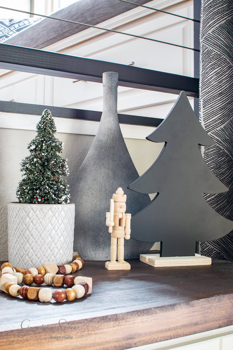Part of the 2020 Christmas house tour, this festive tabletop display showcases a small potted Christmas tree, a wooden nutcracker, a tall gray vase, and a black wooden tree figure. A decorative bead garland elegantly encircles the base of the potted tree.