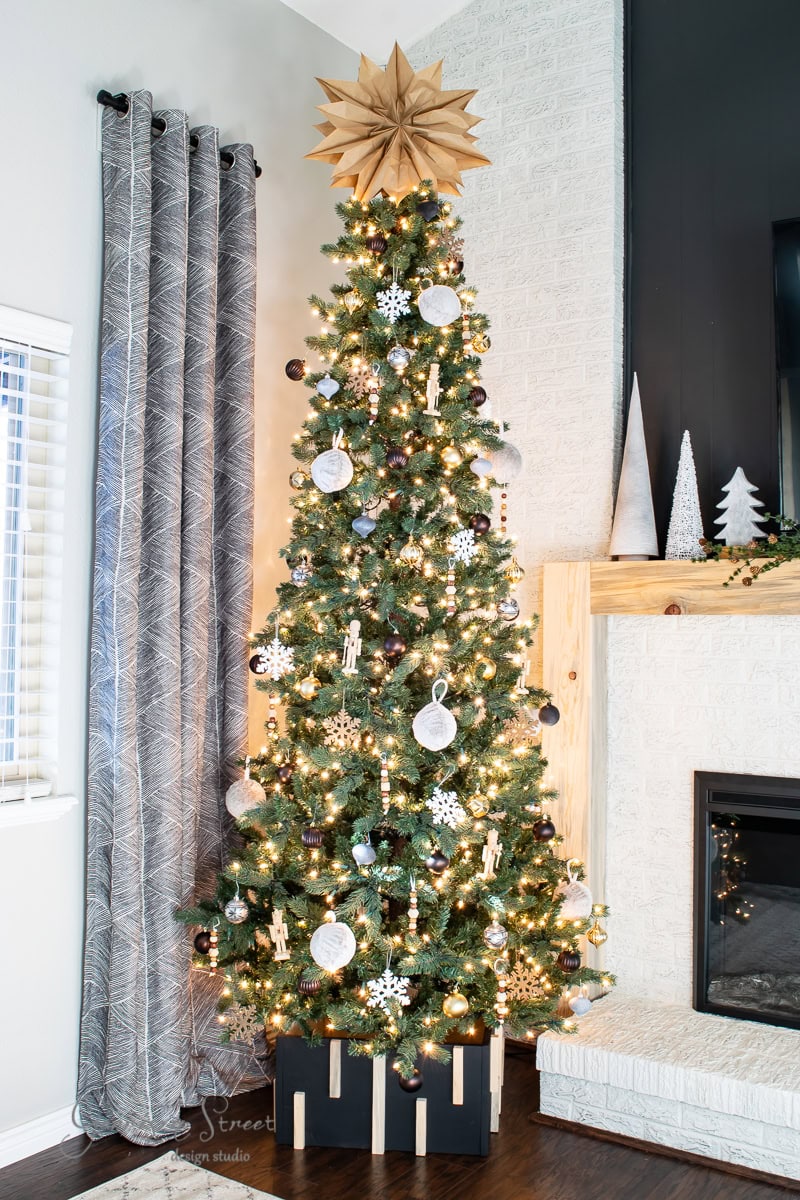 A decorated Christmas tree with a DIY Christmas tree collar stands in the living room next to a fireplace. It is adorned with white lights, snowflakes, baubles, and a large star on top. The room features patterned curtains and a mantel with small decorative trees.