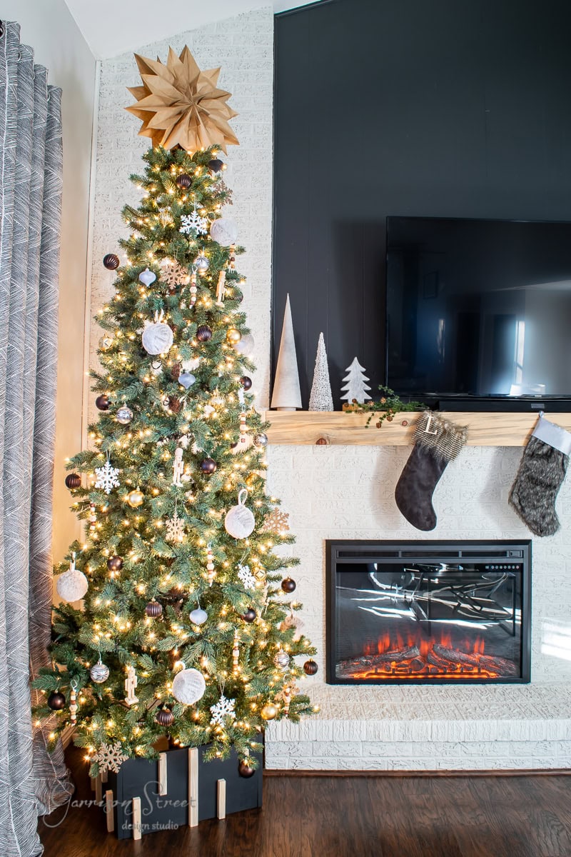 A festive living room showcases a decorated Christmas tree with white and silver ornaments and bright lights. A wooden star tops the tree, which stands elegantly on a DIY Christmas Tree Collar. Nearby, a fireplace with stockings and decorative trees sits beneath a wall-mounted TV.
