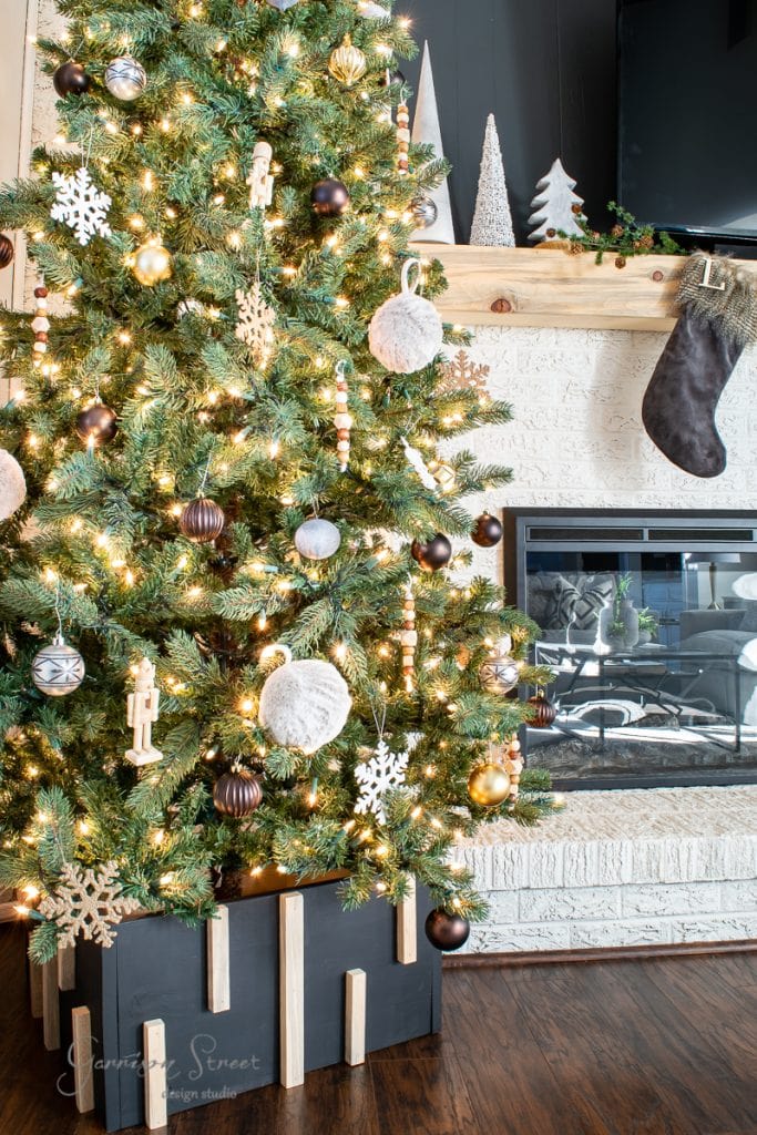 About A decorated Christmas tree with lights and ornaments stands proudly next to a white brick fireplace, adorned with a DIY Christmas tree collar. The mantel showcases festive decorations, including miniature trees and a stocking. The wooden floor enhances the room's warm, cozy ambiance.