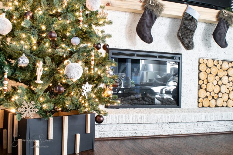 A decorated Christmas tree with lights and ornaments stands beside a modern fireplace, complemented by a DIY Christmas tree collar. Above the fireplace, three stockings hang, marked with the letters L, B, and an unmarked one. Logs are stacked decoratively next to the fireplace.