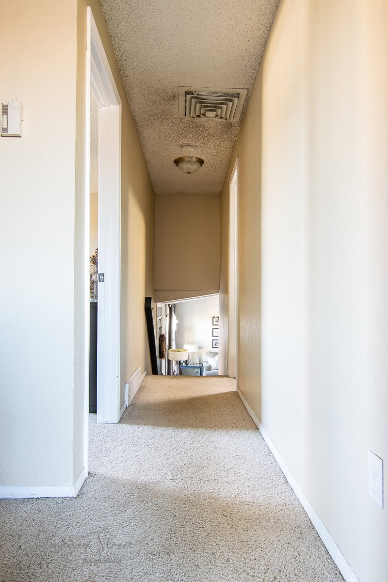 A narrow hallway with beige walls and a carpeted floor reminiscent of 2021 leads to a room. The ceiling features a light fixture and an air vent. Two open doors line the left side, while the end of the hallway offers a glimpse of a room adorned with framed pictures.