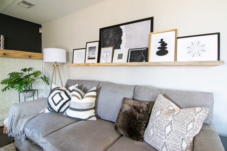 A cozy living room with a gray couch adorned with patterned pillows. Above, discover gallery wall ideas behind the couch where a wooden shelf displays framed artwork in various styles and sizes. A potted plant and a floor lamp are positioned next to the couch.