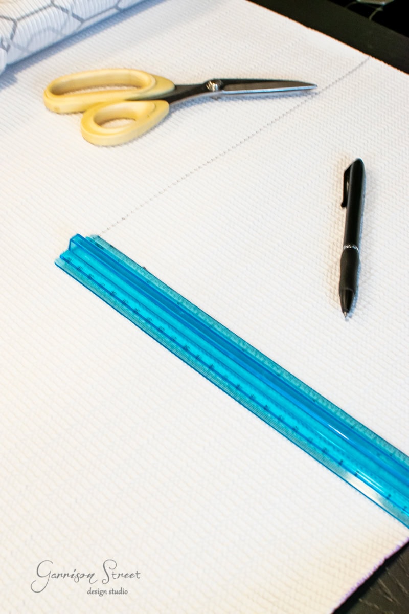 A sheet of white textured fabric is laid out on a surface, demonstrating perfect organization. A pair of scissors, a black pen, and a blue ruler are neatly placed on top. The background includes partial views of other materials that hint at the creativity in this workspace.