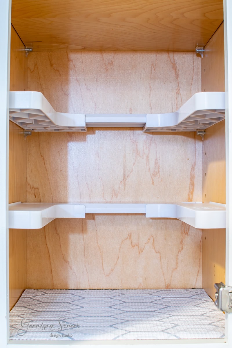 An open wooden cabinet designed for spice organization features two white pull-out shelves and a patterned mat at the bottom. The interior boasts a light wood finish, and the partially visible cabinet door adds an elegant touch on the right side.