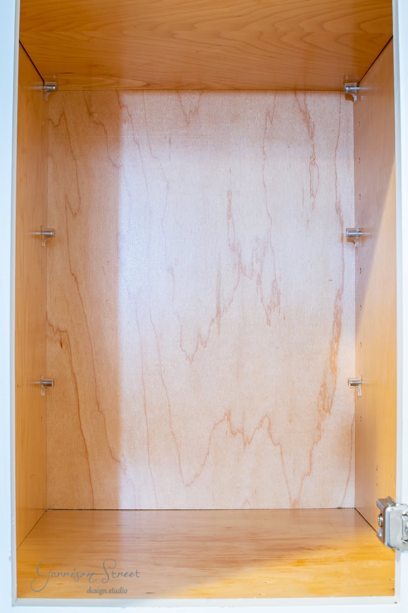 An empty wooden cabinet, perfect for spice organization, features hinges on the sides and a light natural finish. The back panel boasts a distinct wood grain pattern as sunlight softly illuminates its interior.