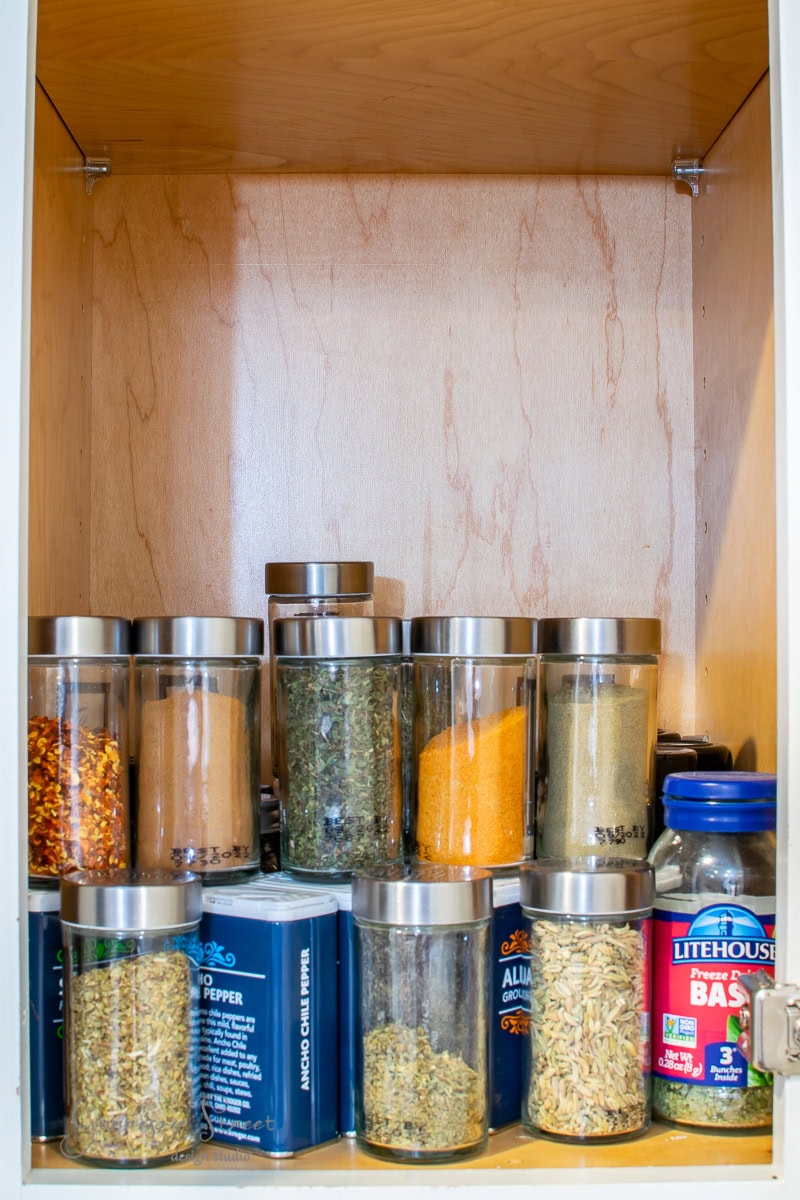 A well-organized kitchen cabinet shelf showcases an array of spice jars. Metal-lidded glass containers brim with colorful spices—orange, yellow, and green. Blue rectangular boxes are neatly labeled, ensuring efficient spice organization.