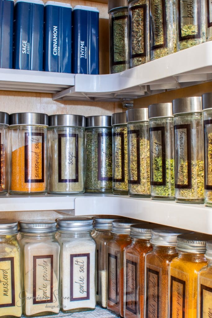 Home An organized spice cabinet features labeled glass jars filled with spices and herbs like sage, cinnamon, thyme, and court bouillon. The jars are neatly arranged in a wooden cupboard, with some spices in bottles and others on a rotating shelf for optimal organization.