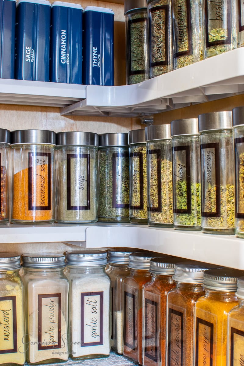 An organized spice cabinet features labeled glass jars filled with spices and herbs like sage, cinnamon, thyme, and court bouillon. The jars are neatly arranged in a wooden cupboard, with some spices in bottles and others on a rotating shelf for optimal organization.