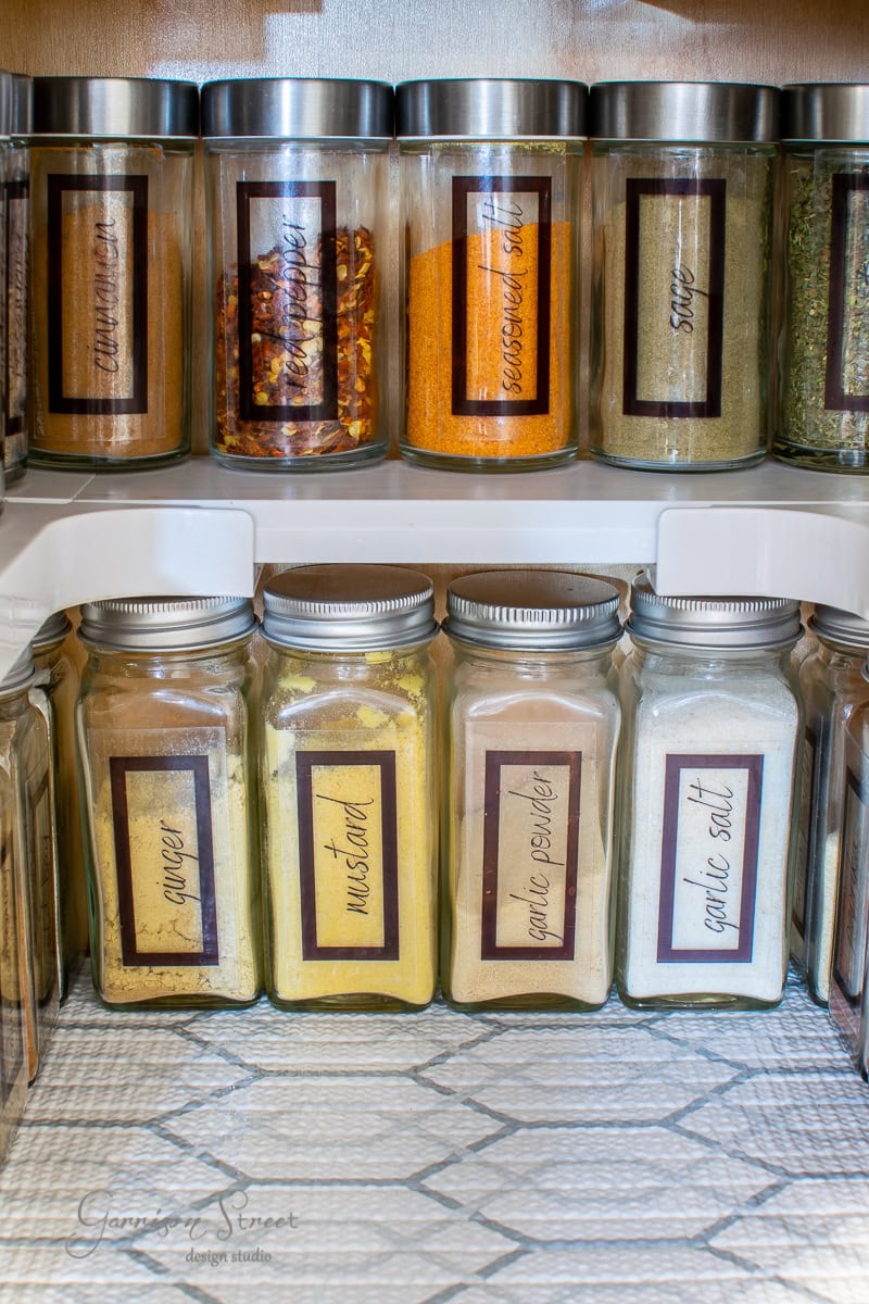 A well-organized kitchen spice cabinet features glass jars filled with vibrant seasonings. The top shelf showcases jars of turmeric and red pepper flakes, while the bottom holds larger, clearly labeled jars of ginger, mustard, and garlic, each with charming handwritten tags.