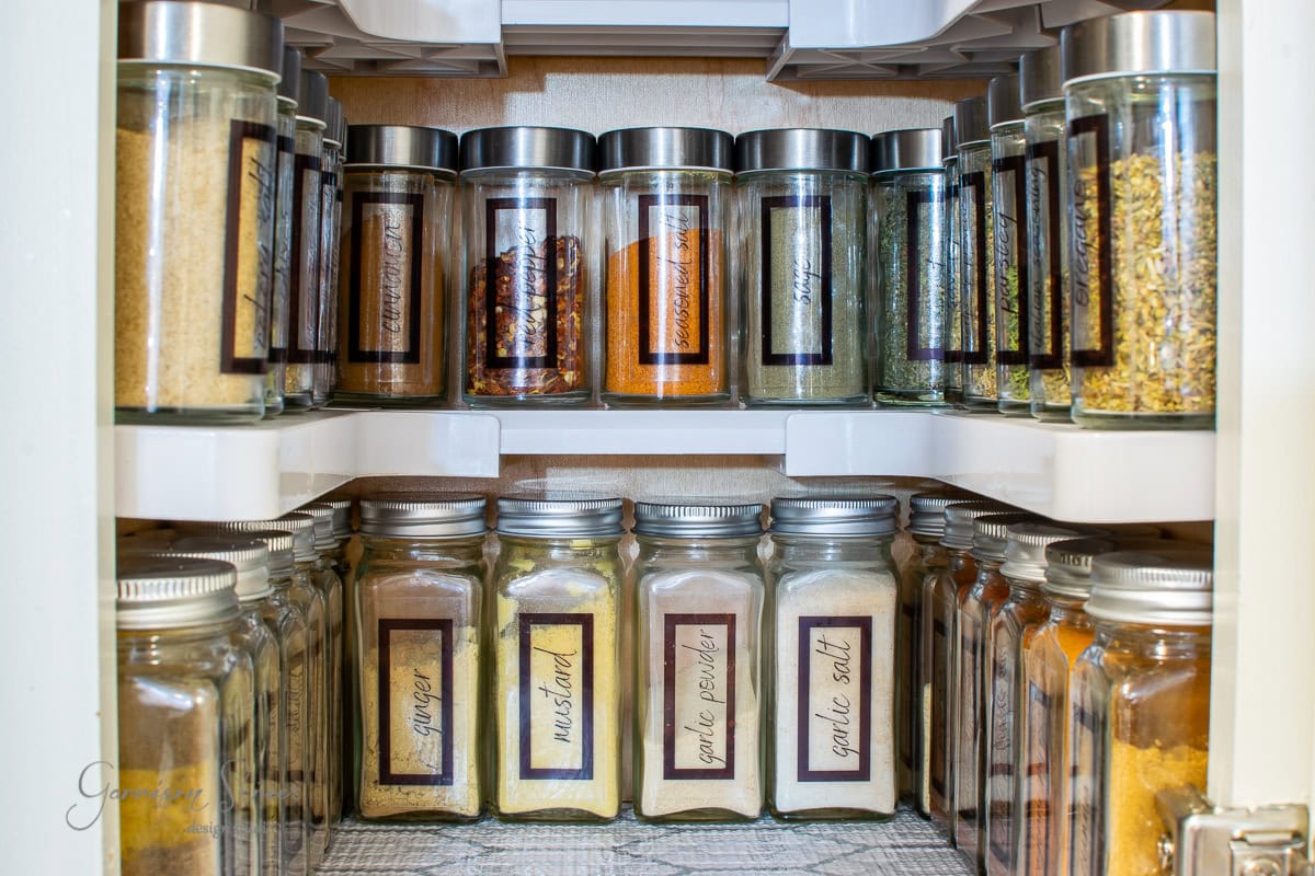 A neatly organized spice cabinet showcases glass jars of various spices. The labeled jars, featuring ginger, mustard, white pepper, and garlic salt, are arranged with precision on rotating shelves for optimal organization.