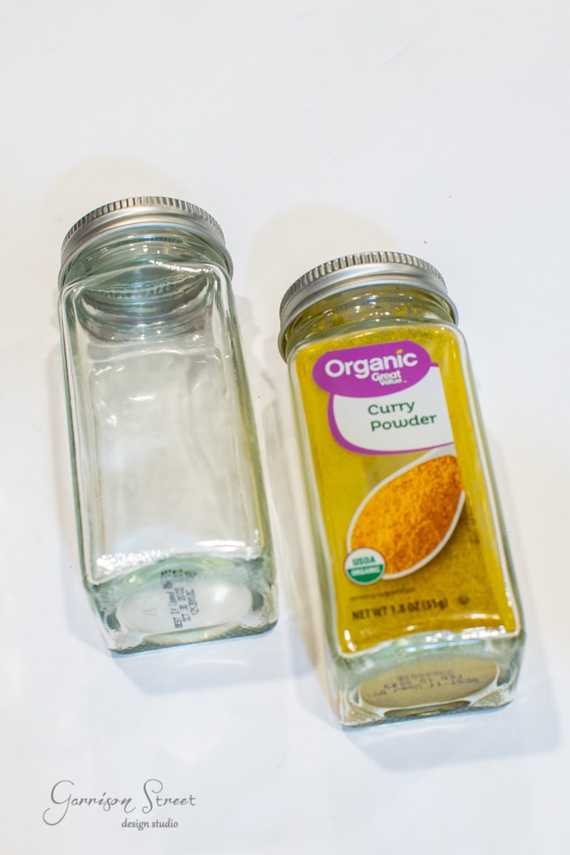 Two glass spice jars sit on a white surface. One jar, perfect for organization in your spice cabinet, is empty and open, while the other is labeled "Organic Curry Powder" and remains sealed.