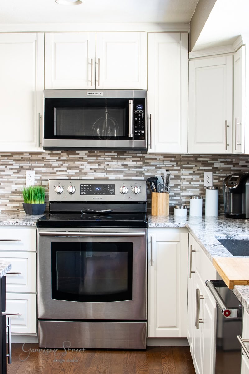 A modern kitchen features stainless steel appliances, including an oven and microwave. White cabinets provide ample storage and surround the appliances, enhancing organization. A mosaic tile backsplash adds texture above the stove, while a utensil holder and small appliances sit on the countertop.
