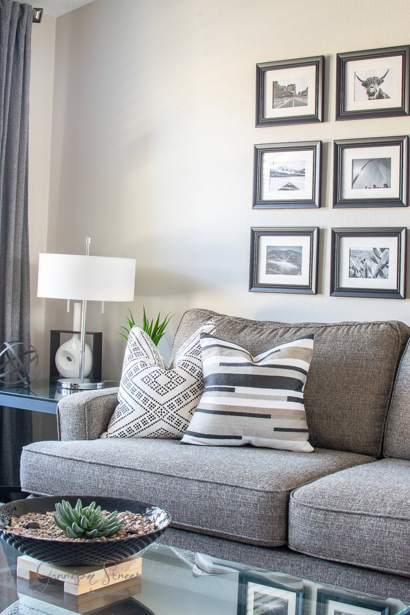 A modern living room features a gray couch with two patterned pillows and an array of DIY art framed in black-and-white on the wall. A white lamp and decorative objects grace the side table, while a glass coffee table displays a charming succulent arrangement.