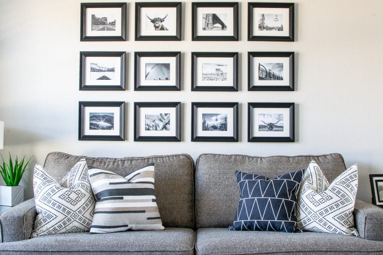 A living room with a gray sofa and patterned pillows in black, white, and gray. Above the sofa, a DIY art display features 15 black-and-white framed photographs arranged in a grid pattern. A small plant in a white pot sits on a side table.