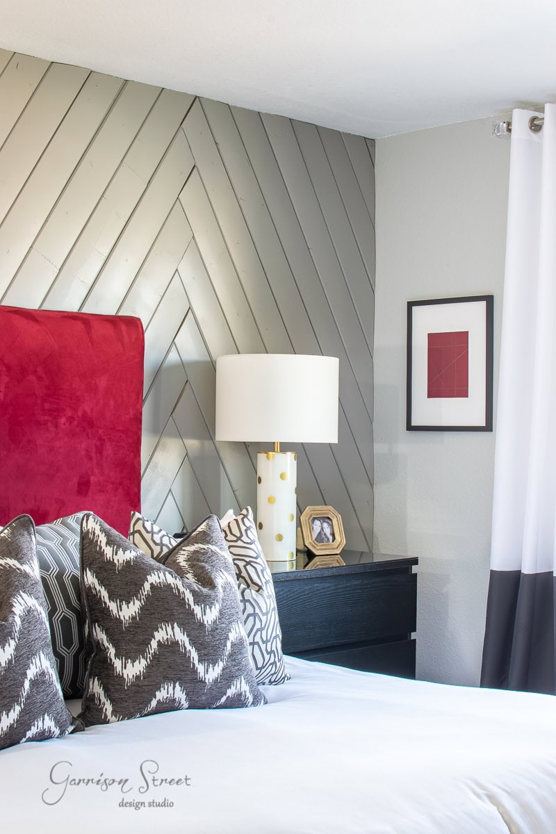 A bedroom features a chevron-patterned gray accent wall and a vibrant red headboard. Patterned pillows adorn the bed, while a white lamp with gold accents rests on the nightstand. A small DIY art piece in a delicate frame adds charm to the space.