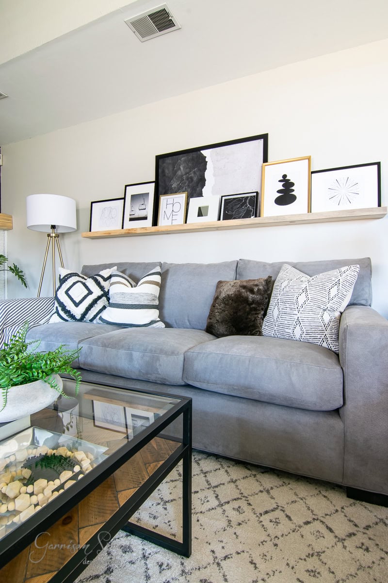 A cozy living room features a gray sofa with patterned pillows, a glass coffee table, and decorative plants. A shelf above displays framed DIY art and photos. The room is softly lit by a floor lamp on the left.