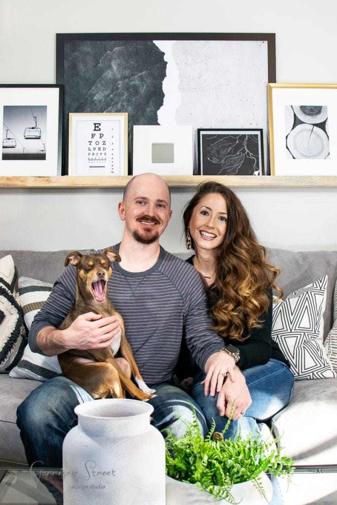 About A smiling couple sits on a couch, with a brown dog on the man's lap. Behind them is a wall shelf with framed art and photos. Decorative pillows and a plant accent the scene. The room is bright and inviting.