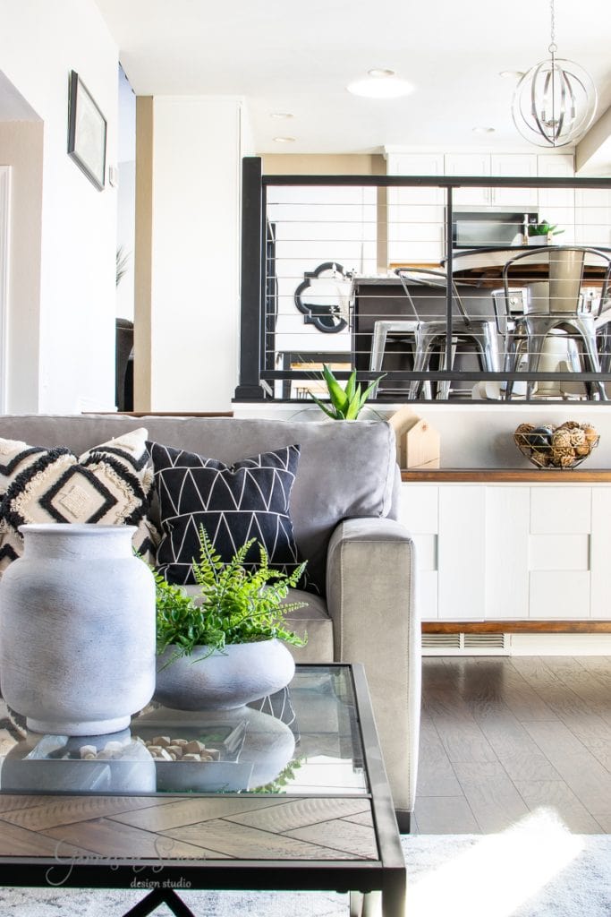 A modern living room with a gray sofa adorned with geometric patterned pillows. A glass coffee table holds decorative vases and plants, showcasing a DIY touch. In the background, a dining area features metal chairs beside cable railing and a spherical pendant light.