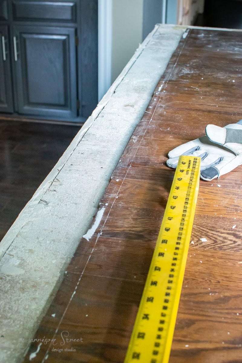 A workbench with a long yellow measuring tape is extended across its surface, perfect for your latest DIY project. A pair of work gloves rests nearby. The wooden bench has a textured edge and a worn appearance, with dark cabinets in the background, ready for your next cable railing installation.