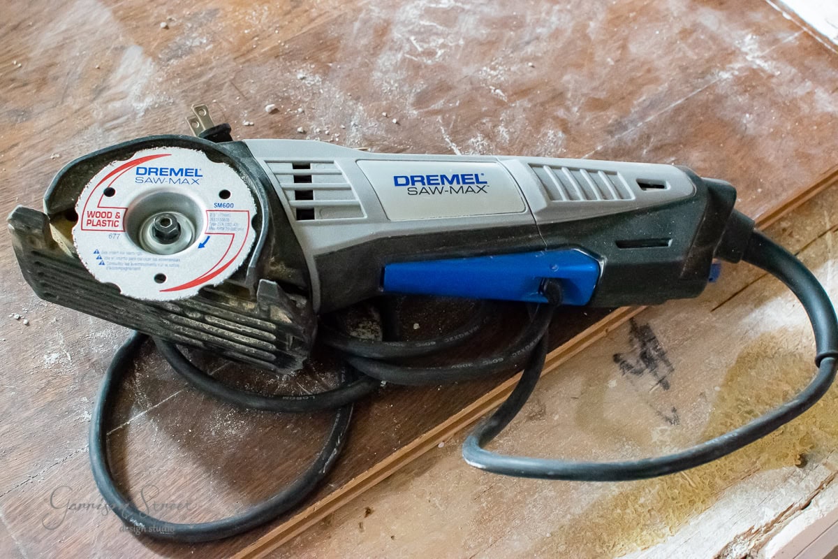 An electric handheld Dremel Saw-Max tool with a circular blade rests on a wooden surface, perfect for your next DIY project. Designed for cutting wood and plastic, it’s an essential tool, whether you’re crafting furniture or installing cable railing. The power cord is neatly coiled beside it.