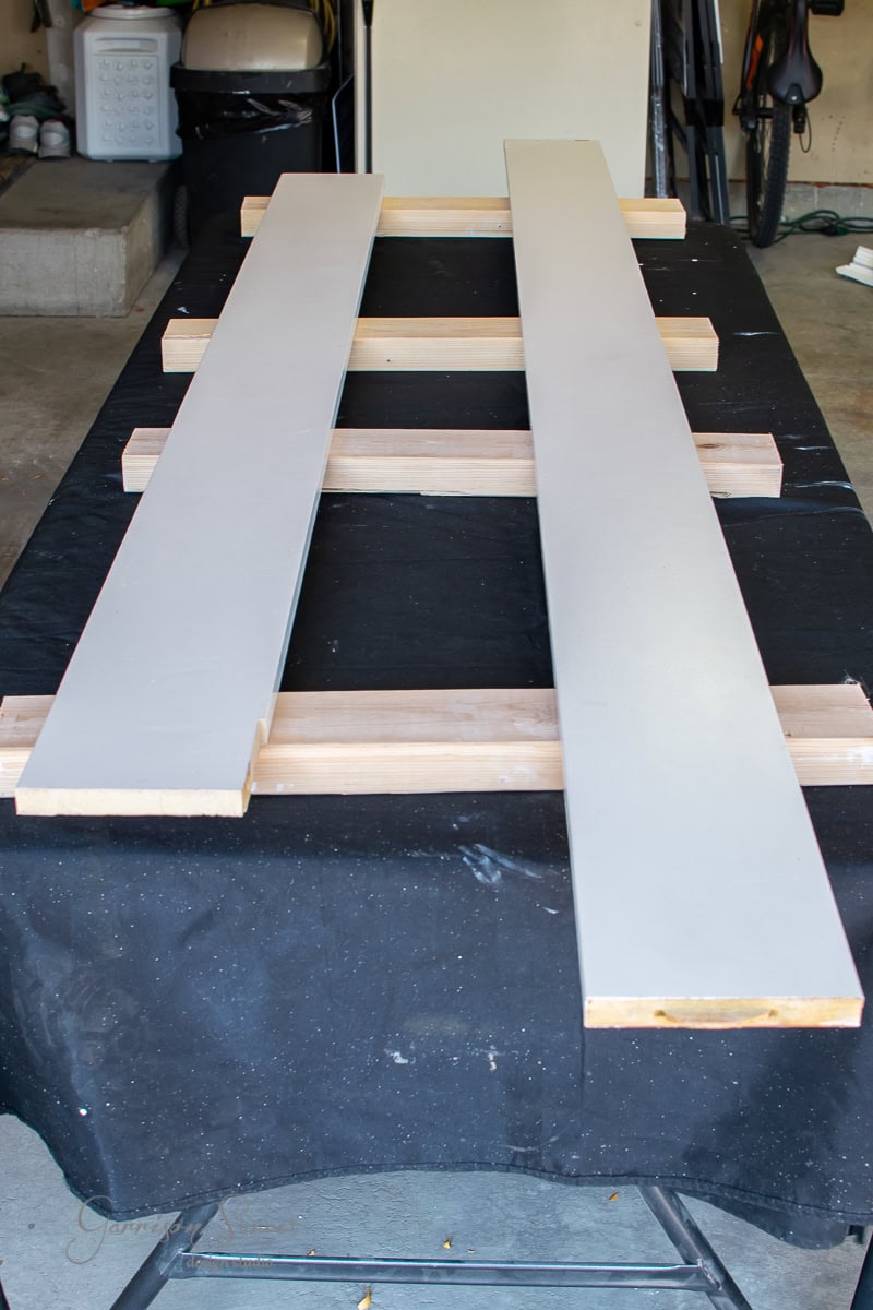 Two long wooden panels painted white are placed parallel on a black surface with wooden supports underneath in a garage setting, showcasing a DIY project. Various garage items and cable railing components are visible in the background.