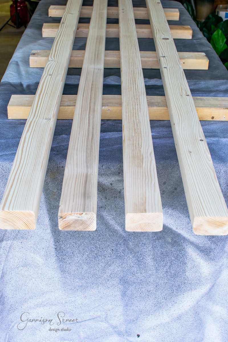 Four wooden planks arranged lengthwise on a gray cloth, supported by smaller horizontal pieces, create a DIY project highlight. The wood has a smooth, unfinished appearance, perfect for incorporating cable railing to infuse modern flair into your handcrafted design.