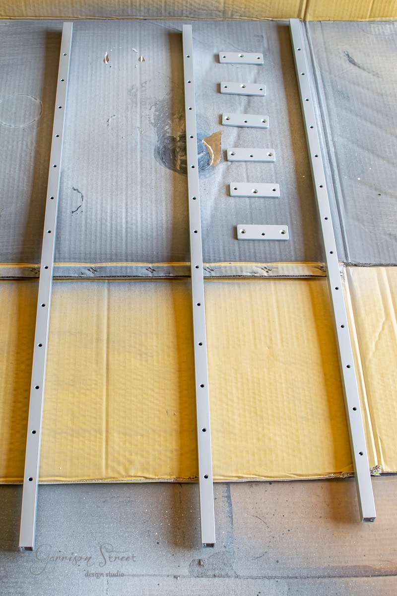 Two long metal bars with evenly spaced holes are laid parallel on a cardboard surface, resembling a DIY cable railing kit. Six small rectangular metal plates are positioned between them, suggesting an arrangement for furniture or shelving assembly.