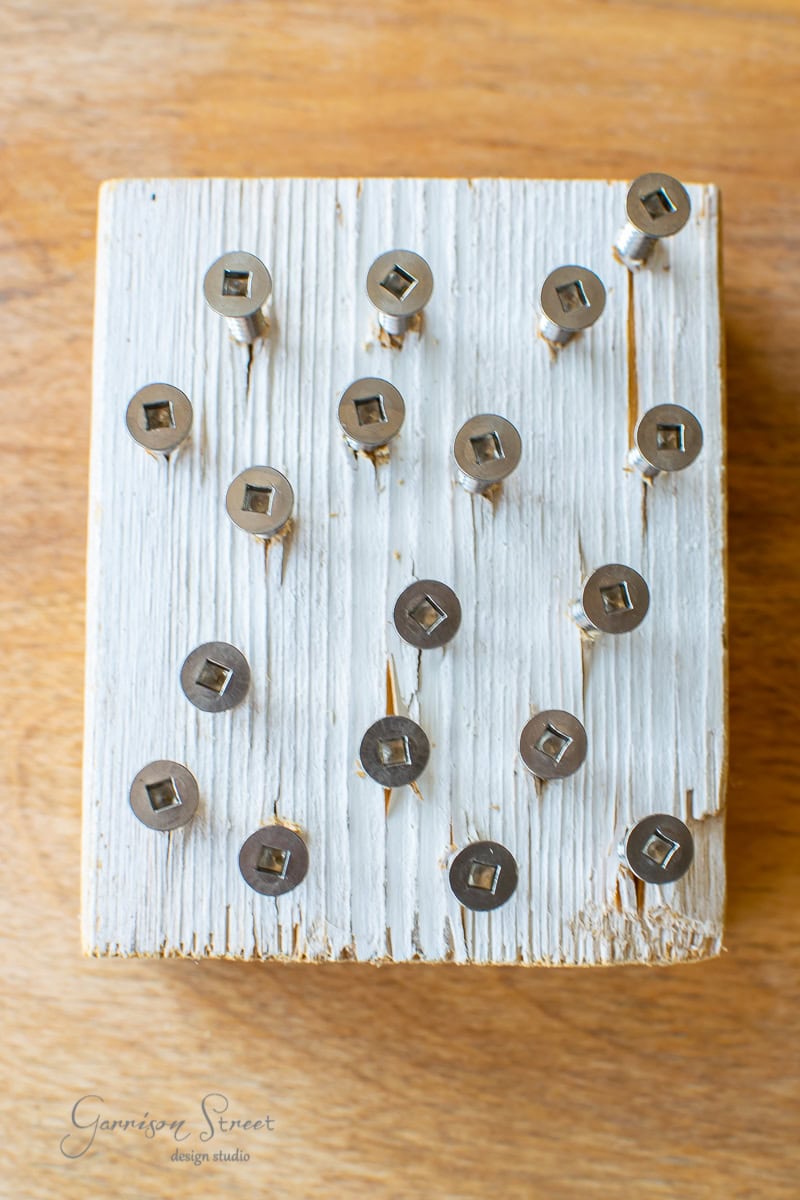 A DIY wooden board with a distressed white finish holds twenty metallic pegs arranged irregularly, reminiscent of a cable railing. The background is a warm, natural wood surface.