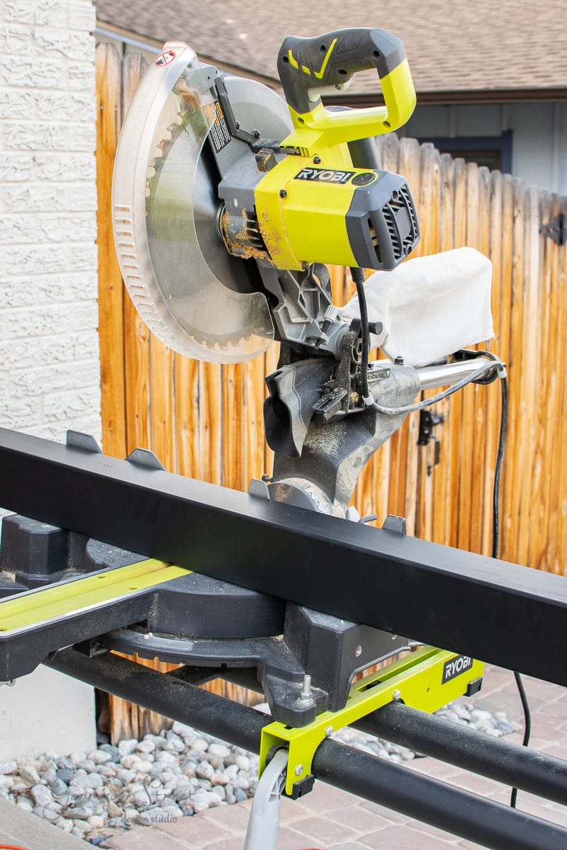 A bright green Ryobi miter saw is positioned on a sturdy black stand outdoors, perfect for any DIY project. Set against a wooden fence and a brick wall, it's ready for precise cutting tasks, whether you're tackling cable railing installations or other woodworking endeavors.