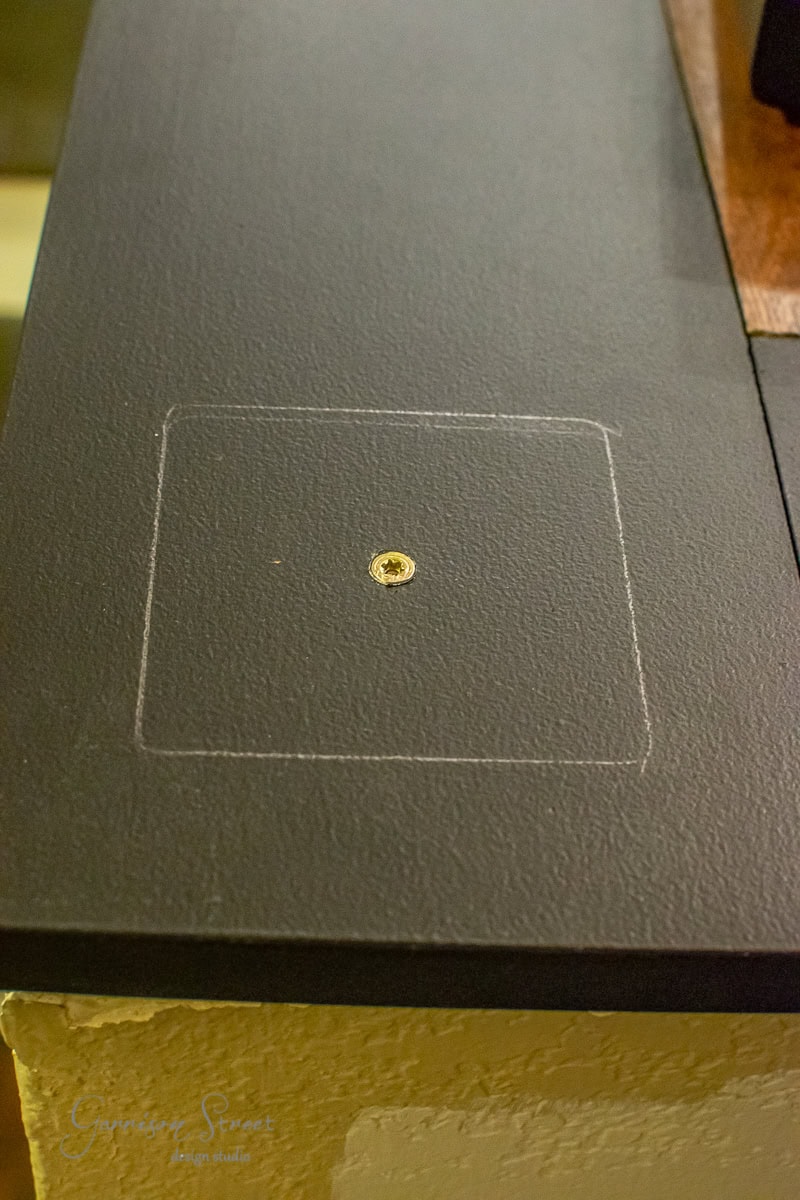 A black surface with a small, gold screw in the center is marked by a faint, hand-drawn square outline, reminiscent of DIY projects. Part of a lighter-colored surface is visible below the black area, suggesting an intricate setup possibly related to cable railing installation.