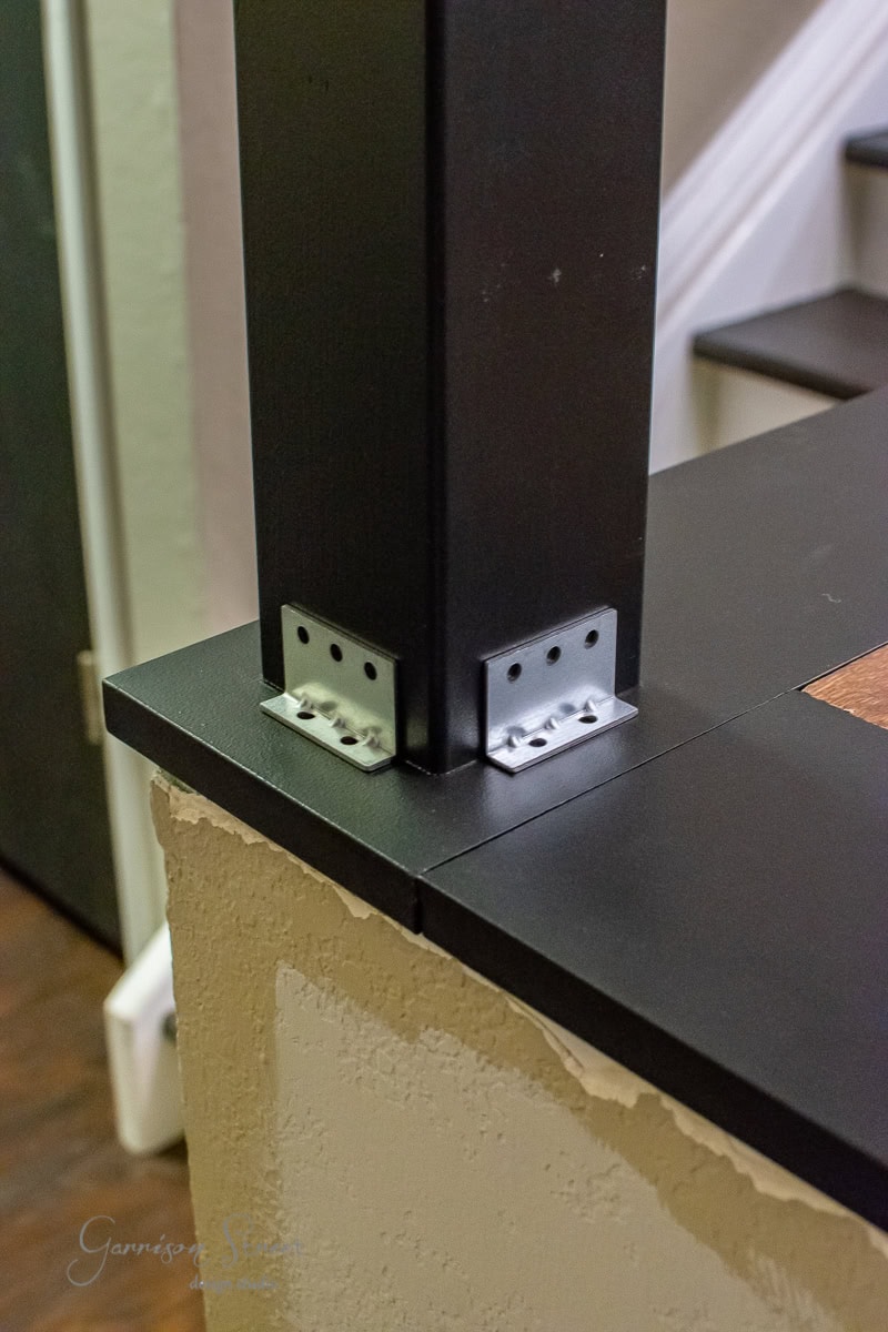 Close-up of a black metal post seamlessly integrated into a DIY cable railing system, attached to the edge of a black surface with two sturdy brackets. The surface rests atop a partially unfinished wall with exposed drywall. In the background, there is a glimpse of an elegant staircase.