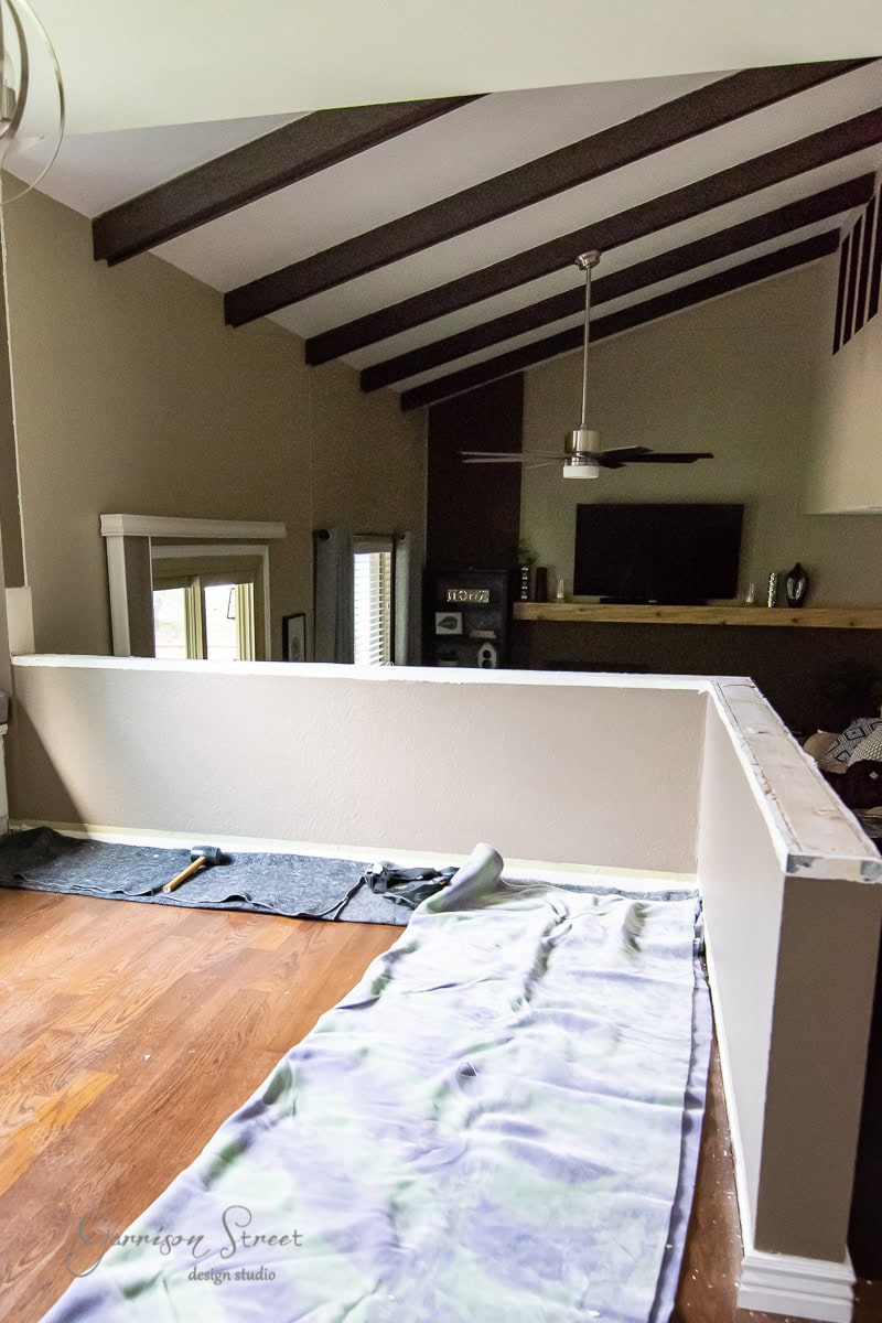 A partially renovated room with a vaulted ceiling and exposed beams showcases a DIY spirit. A low partition wall is under construction, with tarp and tools scattered on the wooden floor. A ceiling fan hums above while windows bring in natural light from the left, highlighting a sleek cable railing design.