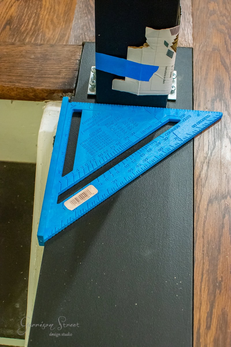 A blue carpenter's square rests on a black surface near a partially painted post, with wood flooring in the background. A label is visible on the tool, hinting at a DIY project involving cable railing. A piece of cardboard is taped to the post, adding to the scene's character.