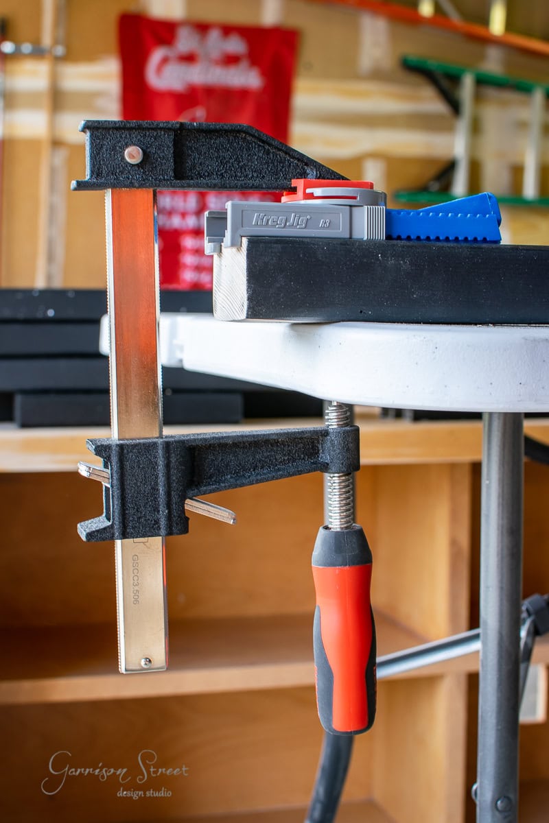 A metal clamp with a red handle secures a tabletop in this well-lit DIY workshop. Boxes are stacked in the background, while a red sign with white text adds to the scene, showcasing tools and cable railing components that complete the workspace.