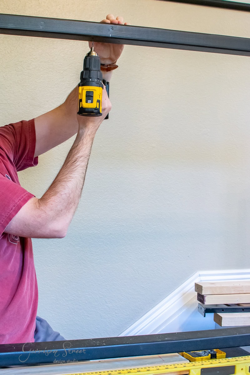 A person in a red shirt is using a power drill on a metal frame, perfecting their cable railing project. The edge of a stair railing and some wooden planks are visible in the background. A yellow spirit level rests at the bottom of the frame, ensuring this DIY endeavor's precision.