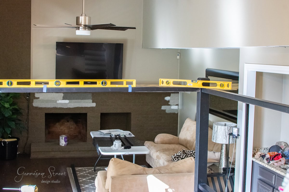 A living room under renovation features a beige couch, a coffee table, and a fireplace with the TV above it. A construction beam with a level tool hints at ongoing DIY projects, while cable railing sections await installation. White paint patches add to the scene's work-in-progress charm.