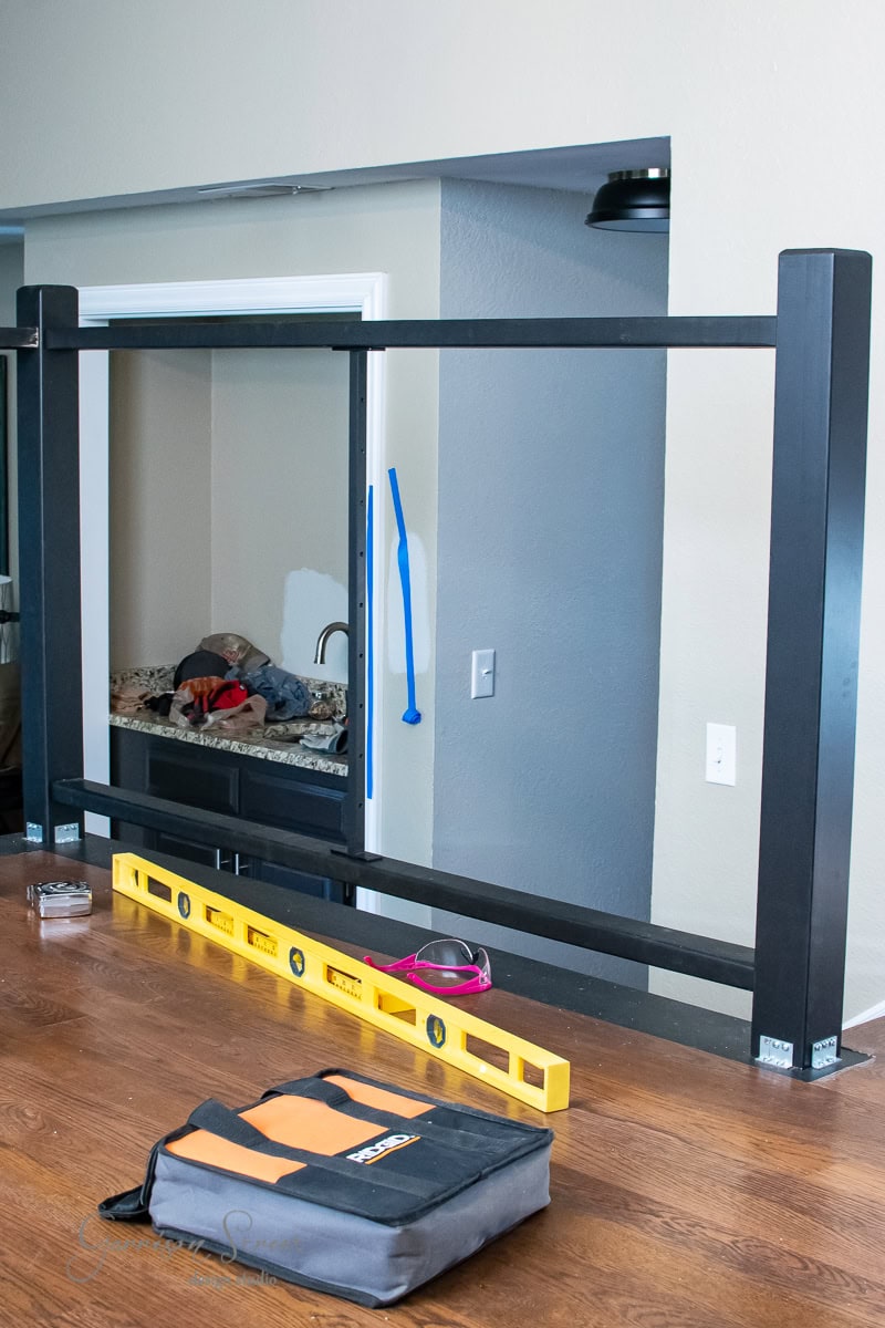 A partially installed black metal cable railing on a wooden floor showcases a DIY project underway, with a yellow level and tool bag nearby. In the background, a blue level leans against the wall, ready to ensure precision in every step.