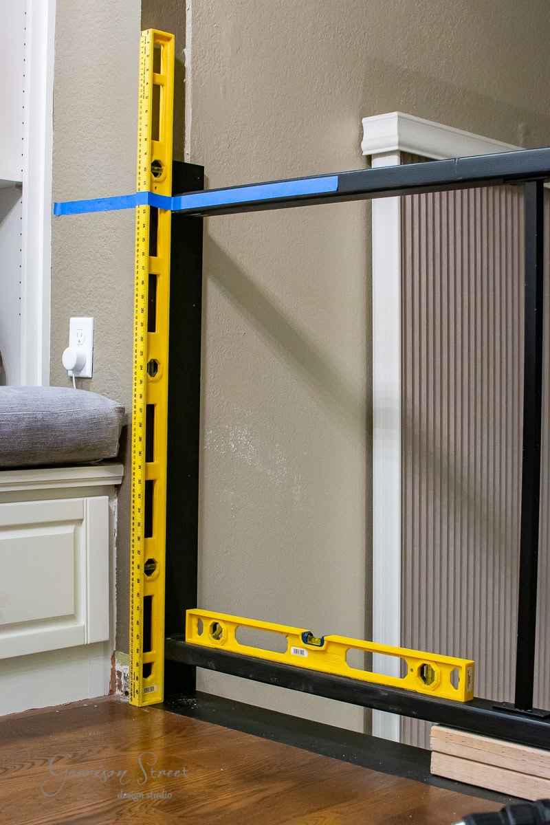 A DIY woodworking setup with a metal frame in progress features a yellow spirit level and carpenter's square ensuring precision. Cable railing components lie nearby, ready for installation. In the background, a white cabinet and power outlet can be seen.