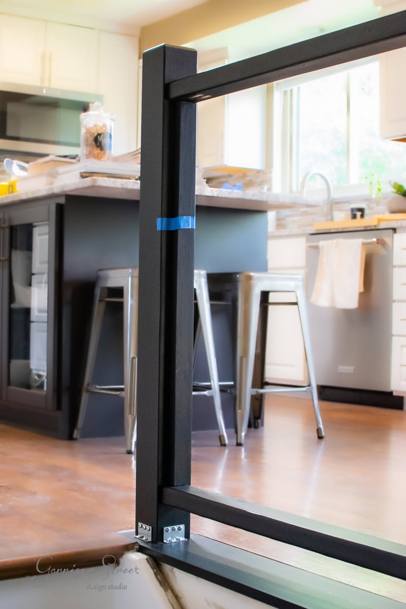A modern kitchen under DIY renovation showcases an unfinished cable railing in the foreground. The sleek space features stainless steel appliances, bar stools at a dark island counter, and large windows that flood the area with natural light.
