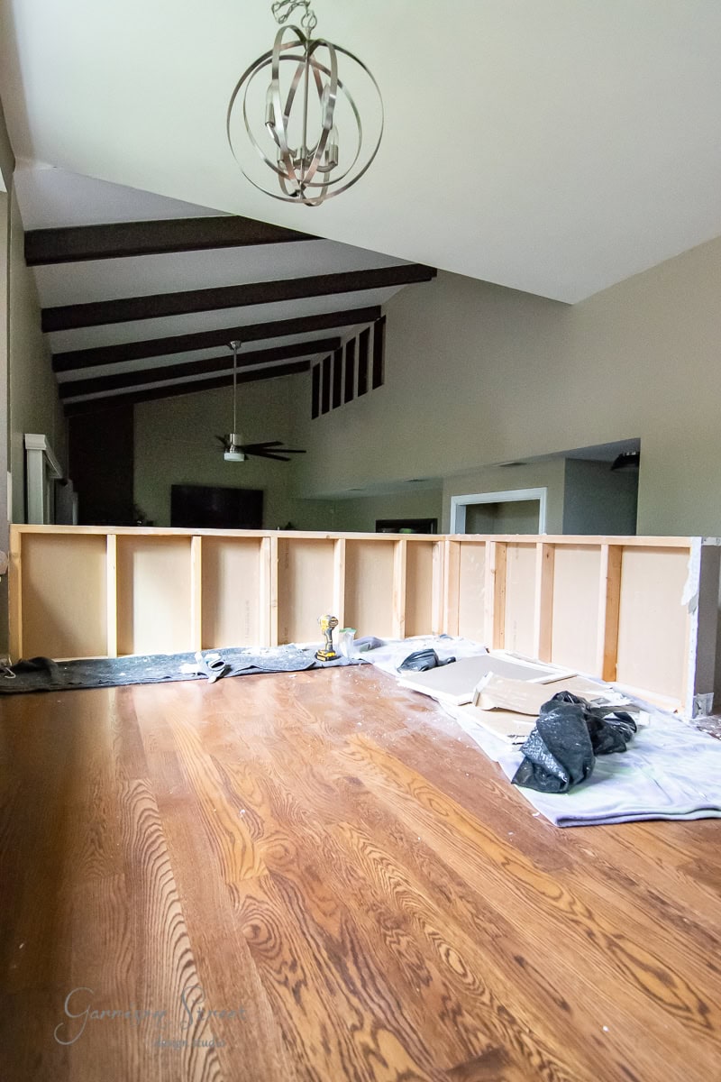 A room under renovation featuring exposed beams and a chandelier, with DIY charm. Unfinished wooden cabinets line the walls, while the floor is littered with tools, tarps, and construction materials. Sunlight filters through a window, highlighting a sleek cable railing in progress.
