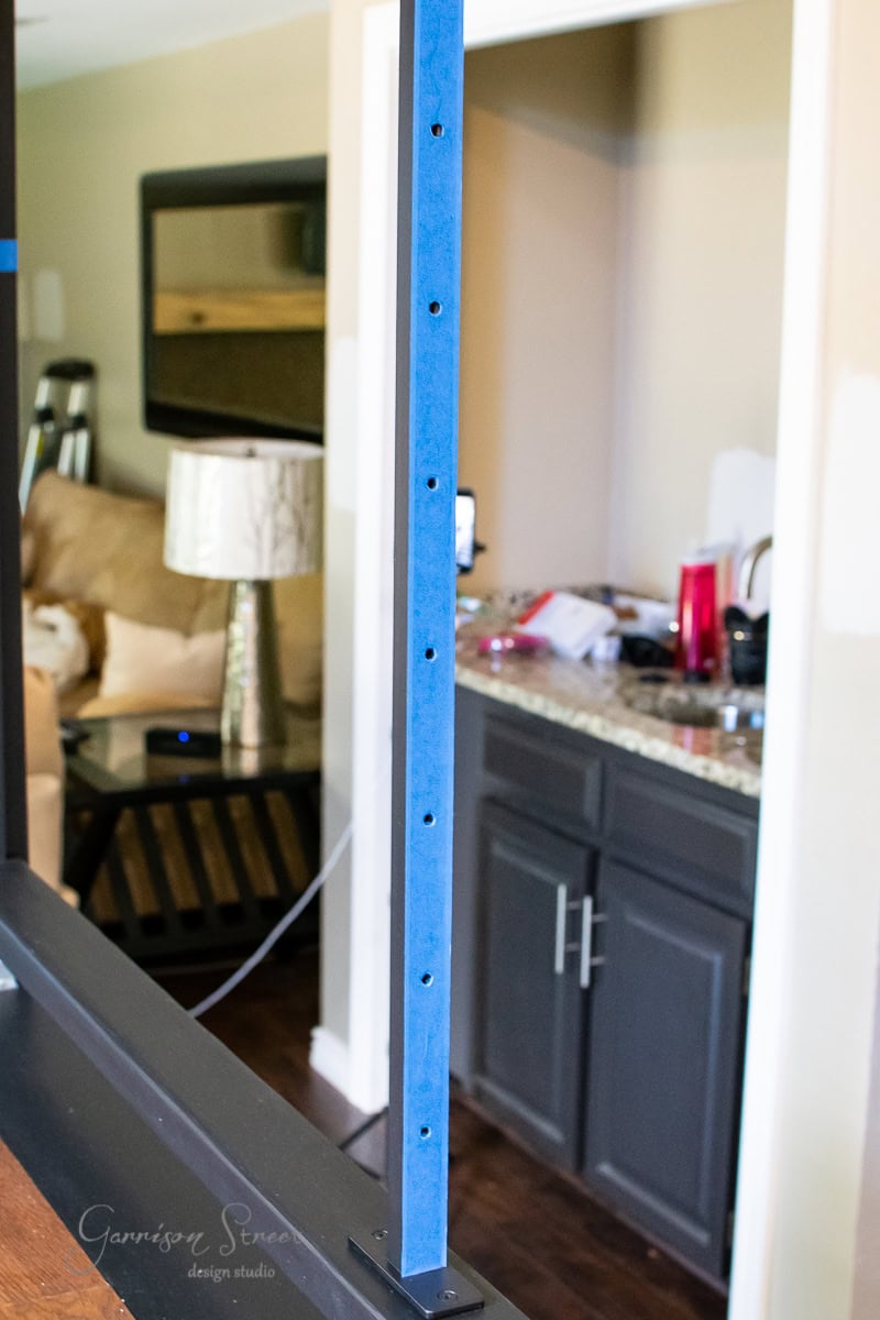 Close-up of a blue metal beam with drilled holes, an essential part of a DIY cable railing project. Behind it, a cozy living space features a beige sofa and a lamp on a side table. To the right, a kitchen counter with cabinets and some kitchenware is visible.