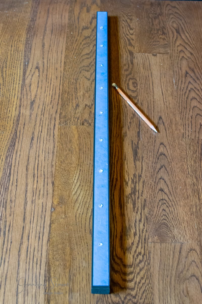 A long blue wooden stick with small screws lined along its center, resembling a cable railing component, is placed on a wooden floor. A sharpened pencil lies parallel to the stick on the right side, suggesting an ongoing DIY project.