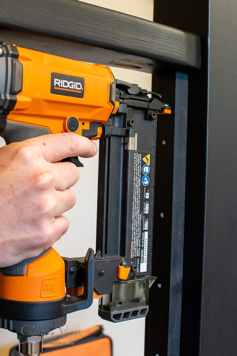 A person skillfully uses an orange Ridgid nail gun on a black frame, showcasing their DIY prowess. The tool is held in the right hand, and various safety labels are clearly visible. This setup could enhance any cable railing project with precision and ease.